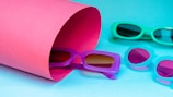 Four pairs of colorful sunglasses are arranged against a vibrant background. A rolled pink sheet partially covers one pair, while the others are positioned on the blue surface.