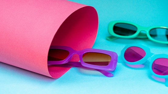 Four pairs of colorful sunglasses are arranged against a vibrant background. A rolled pink sheet partially covers one pair, while the others are positioned on the blue surface.