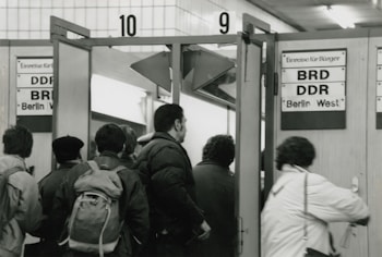 A group of people is standing and walking through a doorway marked with signs indicating entry to different regions, specifically 'BRD' and 'DDR' indicating West and East Berlin. The setting appears to be indoors with tile walls and fluorescent lighting overhead.