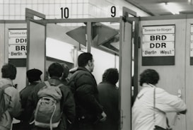 A group of people is standing and walking through a doorway marked with signs indicating entry to different regions, specifically 'BRD' and 'DDR' indicating West and East Berlin. The setting appears to be indoors with tile walls and fluorescent lighting overhead.