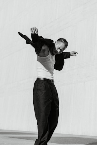 A person with braided hair is actively moving, wearing a white tank top and dark pants, swinging a dark jacket off one shoulder. The background is a plain, lightly textured wall, and the overall ambiance is minimalist with a focus on movement and style.