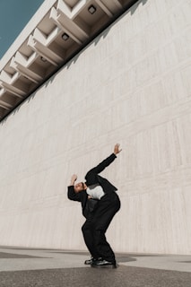 An editorial shot of a man in a modern suit with minimalist accessories, standing against a concrete wall.