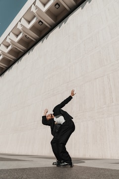 A person dressed in a dark suit and white shirt leans dramatically against a large, textured concrete wall, creating a dynamic pose. The architecture features geometric patterns with a modern and minimalistic design.