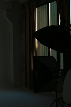 A dimly lit room with photography equipment including a large softbox light placed near a window with closed curtains. The background consists of a plain wall and the area is mostly in shadow.