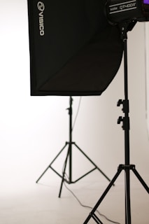 A photo studio setup featuring a black softbox on a stand, with another light stand visible in the background. The scene is minimalistic with clean, white surroundings.