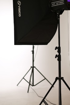 A photo studio setup featuring a black softbox on a stand, with another light stand visible in the background. The scene is minimalistic with clean, white surroundings.