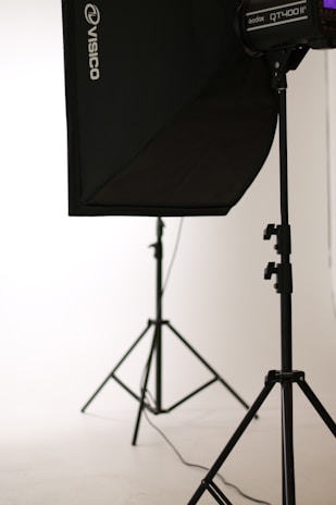 A photo studio setup featuring a black softbox on a stand, with another light stand visible in the background. The scene is minimalistic with clean, white surroundings.