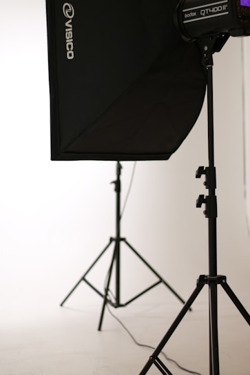 A photo studio setup featuring a black softbox on a stand, with another light stand visible in the background. The scene is minimalistic with clean, white surroundings.