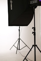 A photo studio setup featuring a black softbox on a stand, with another light stand visible in the background. The scene is minimalistic with clean, white surroundings.