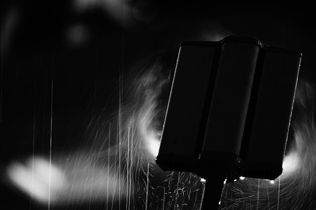 A dark and moody black and white photograph capturing a vertical structure in the foreground, possibly a post or sign, with streaks of rain visible against the background and light creating an atmospheric glow.
