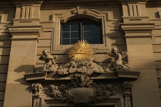 An ornate architectural detail featuring a relief with angelic figures surrounding a golden triangle with an eye at its center, mounted on a classical building facade.