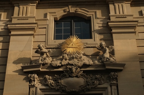 An ornate architectural detail featuring a relief with angelic figures surrounding a golden triangle with an eye at its center, mounted on a classical building facade.