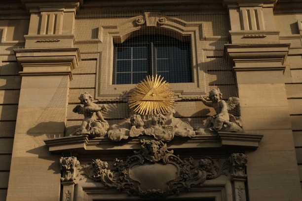 An ornate architectural detail featuring a relief with angelic figures surrounding a golden triangle with an eye at its center, mounted on a classical building facade.