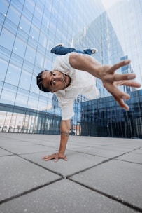 A person is performing a dynamic breakdancing move, balancing on one hand with an outstretched arm and legs in the air. They wear a white, perforated shirt and jeans, while their hair is styled in braids. The setting is an urban environment with a modern glass building in the background, reflecting the surrounding cityscape.