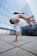 A person is performing a dynamic breakdancing move, balancing on one hand with an outstretched arm and legs in the air. They wear a white, perforated shirt and jeans, while their hair is styled in braids. The setting is an urban environment with a modern glass building in the background, reflecting the surrounding cityscape.