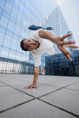 A person is performing a dynamic breakdancing move, balancing on one hand with an outstretched arm and legs in the air. They wear a white, perforated shirt and jeans, while their hair is styled in braids. The setting is an urban environment with a modern glass building in the background, reflecting the surrounding cityscape.