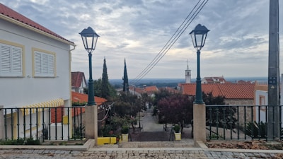 A quaint village scene with a cobblestone pathway lined by trees and traditional houses with red-tiled roofs. Two street lamps frame the path, and a church with a cross on its tower is visible in the background. The sky is overcast with clouds, creating a serene atmosphere.