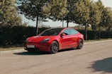 A sleek red electric car is parked on a quiet road surrounded by lush green trees. The car features a polished body, tinted windows, and modern design elements, with a license plate visible at the front.