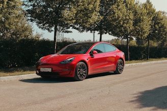 A sleek red electric car is parked on a quiet road surrounded by lush green trees. The car features a polished body, tinted windows, and modern design elements, with a license plate visible at the front.