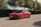 A sleek red electric car is parked on a quiet road surrounded by lush green trees. The car features a polished body, tinted windows, and modern design elements, with a license plate visible at the front.
