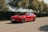 A sleek red electric car is parked on a quiet road surrounded by lush green trees. The car features a polished body, tinted windows, and modern design elements, with a license plate visible at the front.