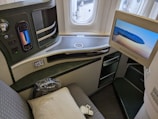 A luxurious airplane seat is shown with an interactive touchscreen control panel, a mounted TV screen displaying an EVA Air airplane, a cushioned seat with a pillow, and various seat controls. The window reveals a view of the airport tarmac.