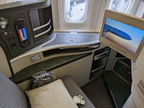 A luxurious airplane seat is shown with an interactive touchscreen control panel, a mounted TV screen displaying an EVA Air airplane, a cushioned seat with a pillow, and various seat controls. The window reveals a view of the airport tarmac.