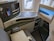 A luxurious airplane seat is shown with an interactive touchscreen control panel, a mounted TV screen displaying an EVA Air airplane, a cushioned seat with a pillow, and various seat controls. The window reveals a view of the airport tarmac.