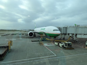 An airplane is parked at the airport gate, connected to the boarding bridge. Ground support vehicles and equipment are present around the plane, which is preparing for boarding or maintenance. The sky is overcast, providing a neutral backdrop.