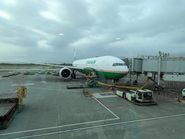 An airplane is parked at the airport gate, connected to the boarding bridge. Ground support vehicles and equipment are present around the plane, which is preparing for boarding or maintenance. The sky is overcast, providing a neutral backdrop.