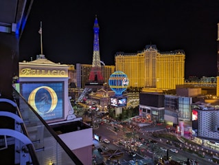 View of a bustling cityscape at night featuring well-illuminated buildings, billboards, and neon lights. Prominent in the foreground is a large Ferris wheel sign and the replica of the Eiffel Tower lit up in red, blue, and white. On the left, there is a large 'Bellagio' sign with a banner for 'O' by Cirque du Soleil. The streets are busy with traffic and pedestrian activity.