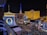 View of a bustling cityscape at night featuring well-illuminated buildings, billboards, and neon lights. Prominent in the foreground is a large Ferris wheel sign and the replica of the Eiffel Tower lit up in red, blue, and white. On the left, there is a large 'Bellagio' sign with a banner for 'O' by Cirque du Soleil. The streets are busy with traffic and pedestrian activity.