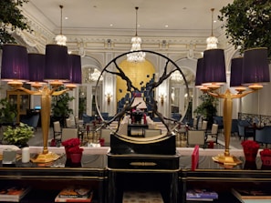 An elegant interior with a sophisticated and opulent design. The room features large chandeliers, ornate moldings, and a prominent decorative piece in the form of a circular sculpture. Purple lampshades provide a warm glow, accentuating the luxurious decor. The furniture includes plush chairs and tables, some adorned with vibrant red floral arrangements. Large mirrors and lush green plants add to the ambient richness of the setting.