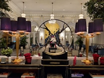 An elegant interior with a sophisticated and opulent design. The room features large chandeliers, ornate moldings, and a prominent decorative piece in the form of a circular sculpture. Purple lampshades provide a warm glow, accentuating the luxurious decor. The furniture includes plush chairs and tables, some adorned with vibrant red floral arrangements. Large mirrors and lush green plants add to the ambient richness of the setting.