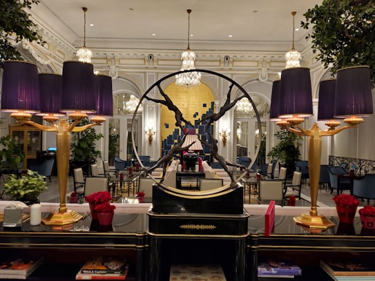 An elegant interior with a sophisticated and opulent design. The room features large chandeliers, ornate moldings, and a prominent decorative piece in the form of a circular sculpture. Purple lampshades provide a warm glow, accentuating the luxurious decor. The furniture includes plush chairs and tables, some adorned with vibrant red floral arrangements. Large mirrors and lush green plants add to the ambient richness of the setting.