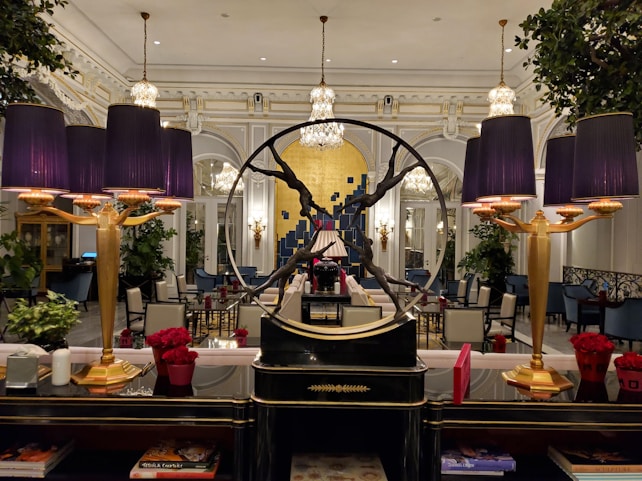 An elegant interior with a sophisticated and opulent design. The room features large chandeliers, ornate moldings, and a prominent decorative piece in the form of a circular sculpture. Purple lampshades provide a warm glow, accentuating the luxurious decor. The furniture includes plush chairs and tables, some adorned with vibrant red floral arrangements. Large mirrors and lush green plants add to the ambient richness of the setting.