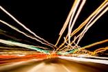 Long exposure photograph capturing light trails of various colors against a dark background. The streaks of light create a dynamic and energetic pattern, suggesting motion and speed.