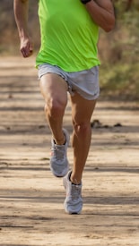 A person is running on a dirt path, wearing a bright green shirt and light gray shorts. Their shoes are athletic and suitable for jogging.