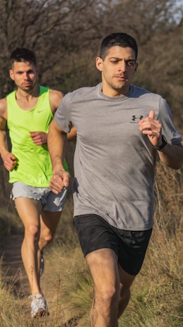 Two men are running on a dirt trail surrounded by dry grass and sparse trees. The man in the foreground is wearing a gray t-shirt and black shorts, while the man behind him is wearing a bright green tank top and light-colored shorts. The sunlight casts clear shadows, indicating a bright and possibly warm day.