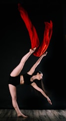A dancer is captured in a dynamic pose, balancing on one foot with her other leg extended high in the air. She is grasping vivid red fabric that flows gracefully behind her. She wears a black athletic outfit and stands against a dark background, enhancing the contrast with the bright red fabric.