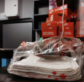 A stack of white napkins is wrapped in a transparent plastic bag on a dark surface. In the background, there are several stacked orange boxes with recognizable branding and a black monitor screen.