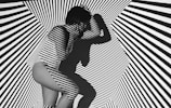 A person is crouched in a space filled with bold black and white diagonal stripes that create an optical illusion effect. The pattern is projected onto the person's body, making them blend into the background. The person's shadow adds another layer of complexity to the visual.