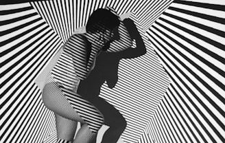 A person is crouched in a space filled with bold black and white diagonal stripes that create an optical illusion effect. The pattern is projected onto the person's body, making them blend into the background. The person's shadow adds another layer of complexity to the visual.