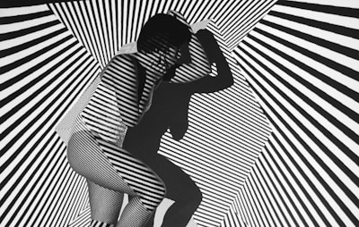 A person is crouched in a space filled with bold black and white diagonal stripes that create an optical illusion effect. The pattern is projected onto the person's body, making them blend into the background. The person's shadow adds another layer of complexity to the visual.
