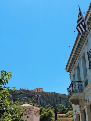 A historic building with classical architecture is situated on a hill. A modern building with balconies and a Greek flag hangs prominently in the foreground. Lush greenery contrasts with a clear blue sky, enhancing the scenic view.