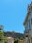 A historic building with classical architecture is situated on a hill. A modern building with balconies and a Greek flag hangs prominently in the foreground. Lush greenery contrasts with a clear blue sky, enhancing the scenic view.