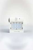 A square white LED light fixture featuring twelve small illuminated LED bulbs arranged in a grid. The fixture has a white casing with a small, round motion sensor above the LED grid and a branding label at the bottom.