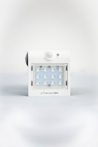 A square white LED light fixture featuring twelve small illuminated LED bulbs arranged in a grid. The fixture has a white casing with a small, round motion sensor above the LED grid and a branding label at the bottom.