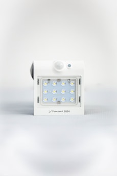 A square white LED light fixture featuring twelve small illuminated LED bulbs arranged in a grid. The fixture has a white casing with a small, round motion sensor above the LED grid and a branding label at the bottom.