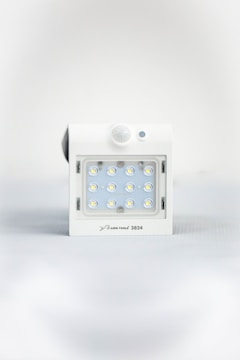 A square white LED light fixture featuring twelve small illuminated LED bulbs arranged in a grid. The fixture has a white casing with a small, round motion sensor above the LED grid and a branding label at the bottom.